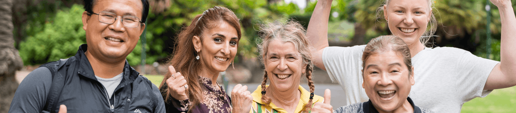 A diverse group of five people outdoors smile and give thumbs-up gestures, with greenery and trees in the background.